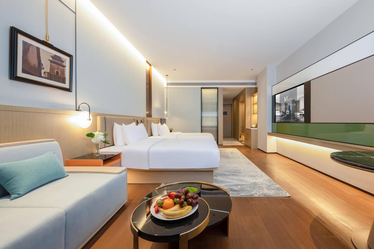 Una habitación de hotel moderna con cama, sofá, una mesa de comedor con fruta y un ventanal grande.
