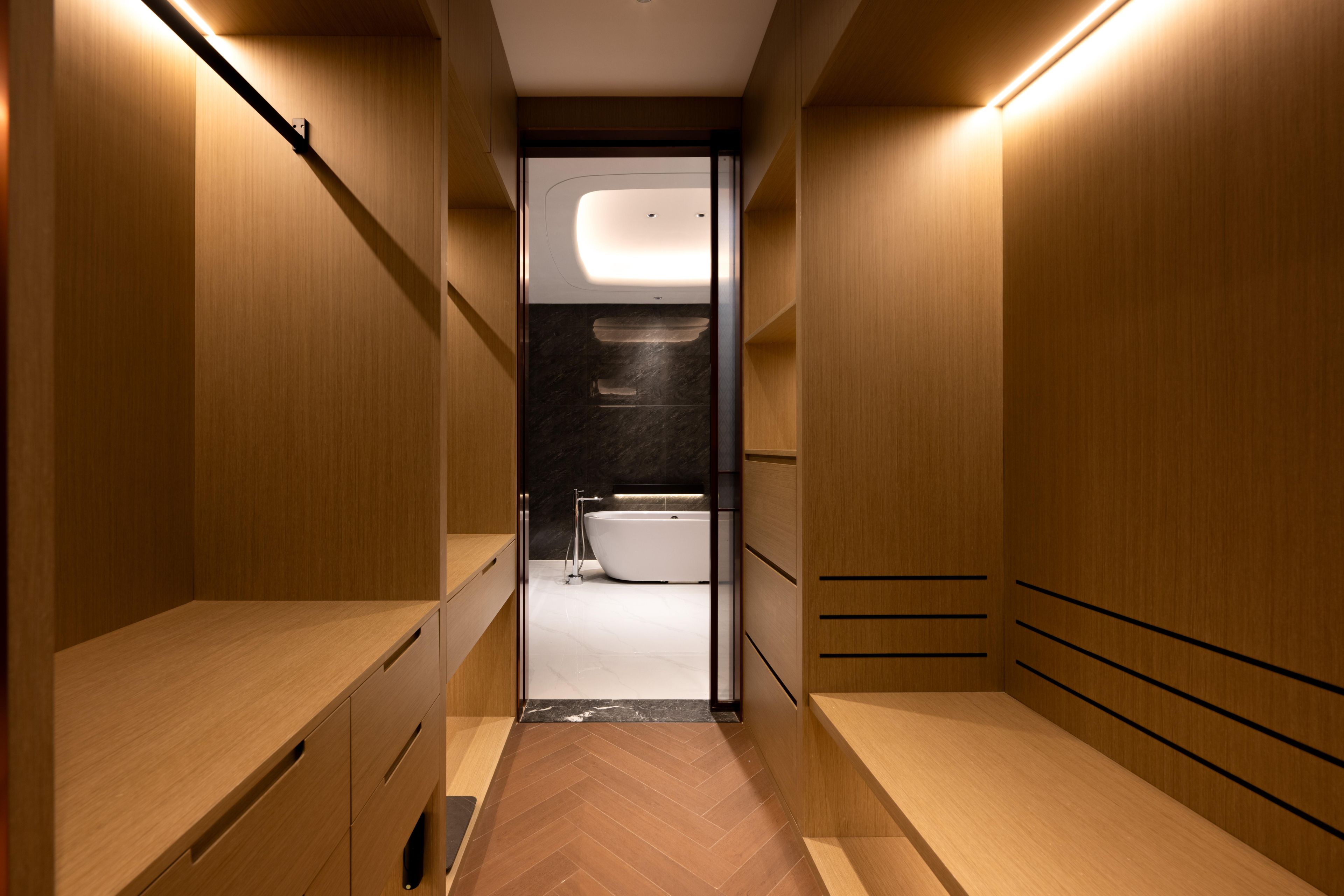 A narrow, wooden-paneled hallway leading to a bathroom with a white bathtub and a modern light fixture.