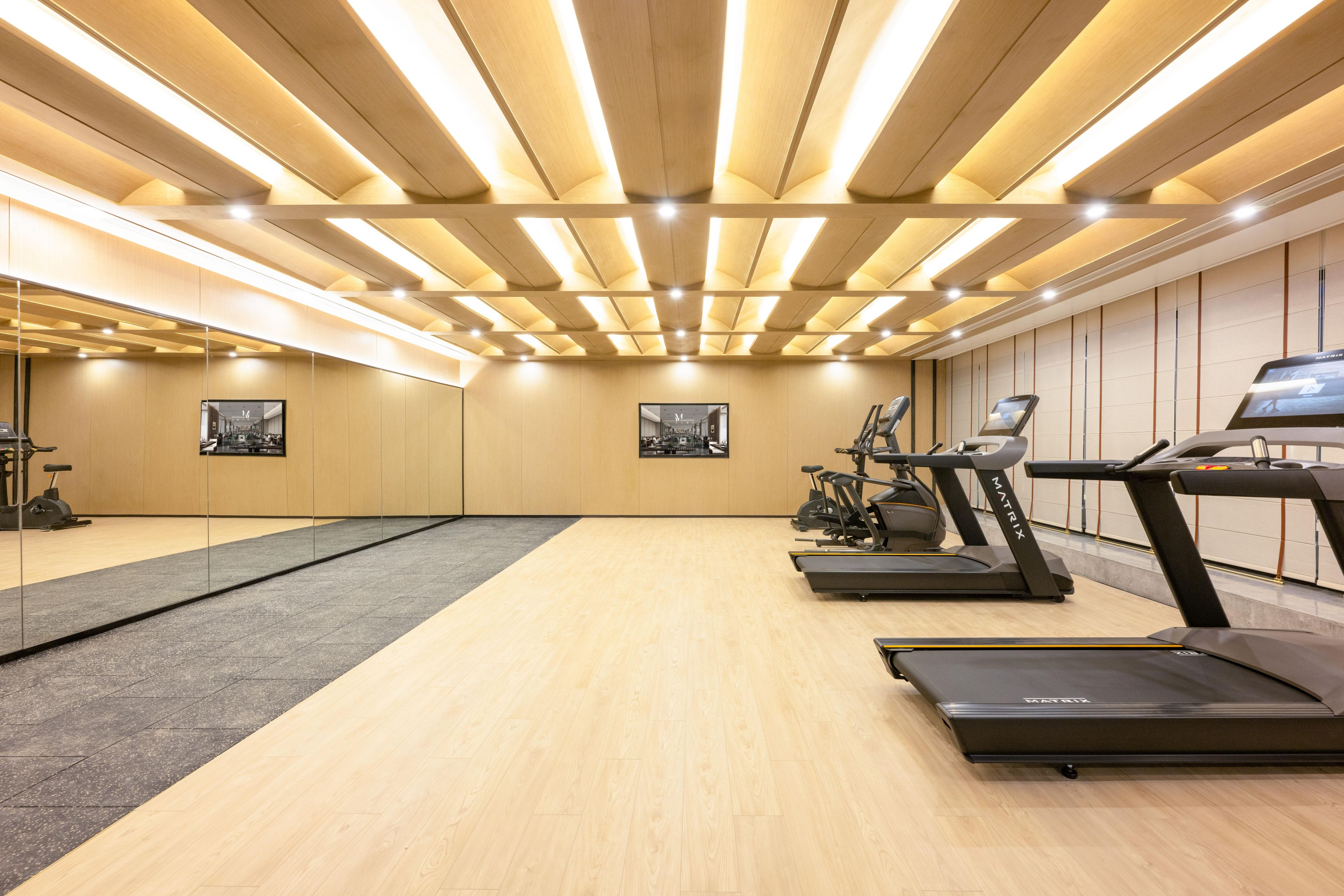 A modern gym with wooden floors, exercise equipment, and a ceiling with linear lighting.