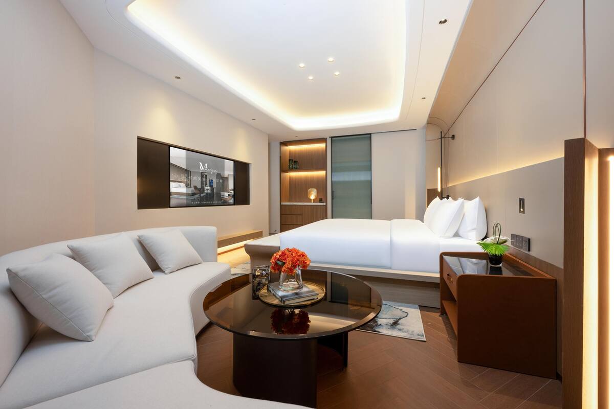 Una habitación de hotel moderna con un sofá, una cama, una mesa de centro de cristal con un florero y una mesita de noche con una lámpara.
