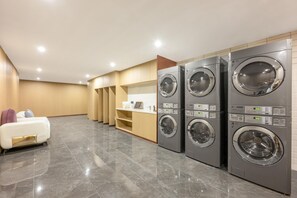 Un lavadero con tres lavadoras de carga frontal apiladas, un sofá blanco y un estante de madera con diversos objetos.