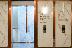 Interior de un ascensor con paredes y suelo de mármol, pantalla digital que muestra los números de los pisos, y rótulos para habitaciones de huéspedes y salas de reuniones.