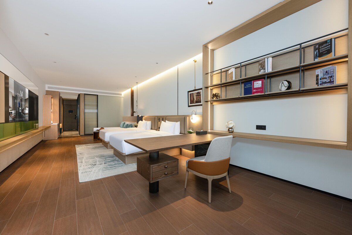 Una habitación de hotel moderna con una cama grande, un escritorio, una silla y una estantería con libros y un reloj.