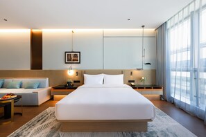 Una habitación de hotel moderna con una cama grande, un sofá, una mesa de comedor con fruta y vistas a la ciudad a través de grandes ventanales.