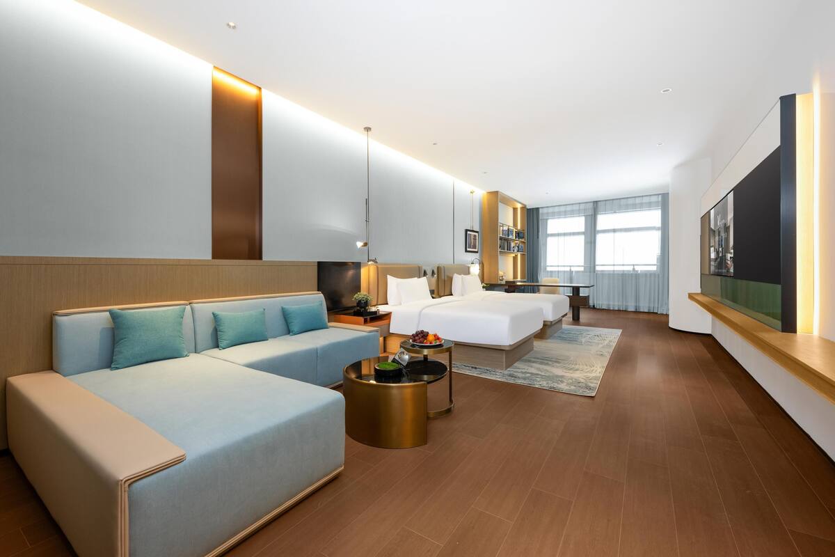 Una habitación de hotel moderna con un sofá, dos camas individuales, un televisor y una pequeña mesa con fruta.
