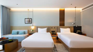 A modern hotel room with two single beds, a sofa, a desk, and a dining area.
