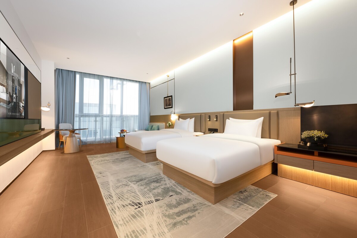 A modern hotel room with two beds, a large window with curtains, a flat-screen TV, and a wooden floor.