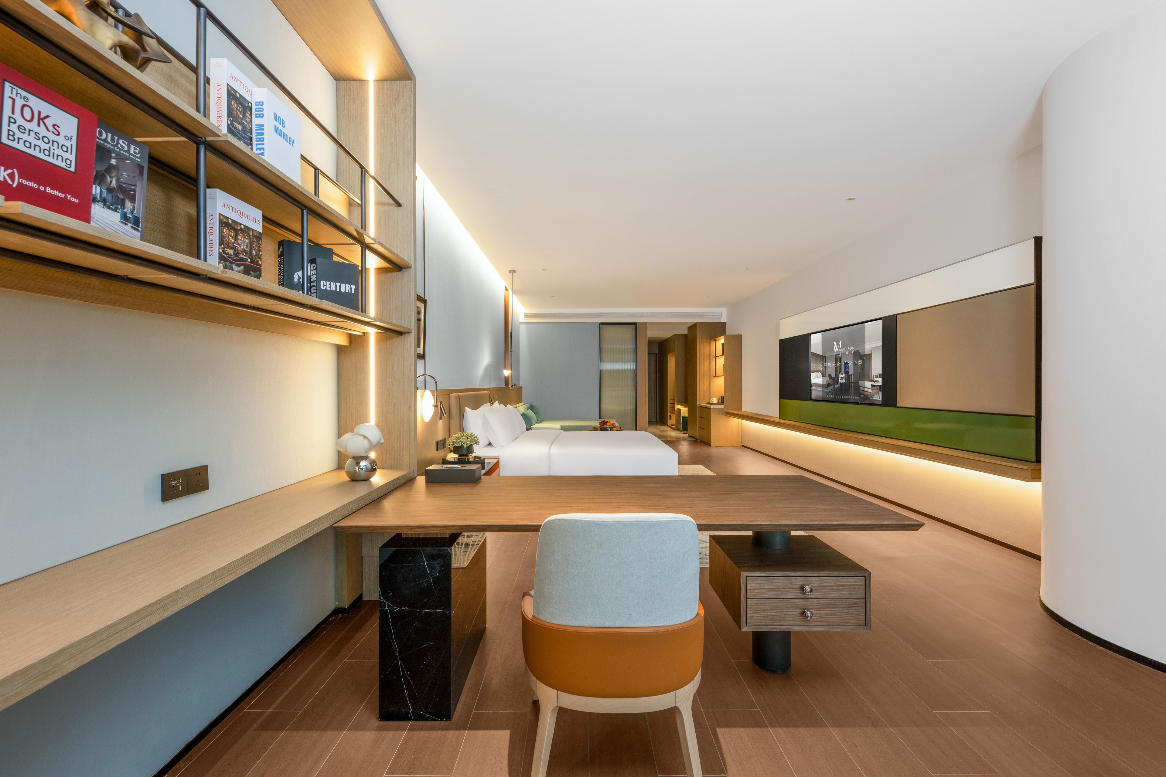 A modern hotel room with a large bed, a desk, a chair, and a bookshelf with books.