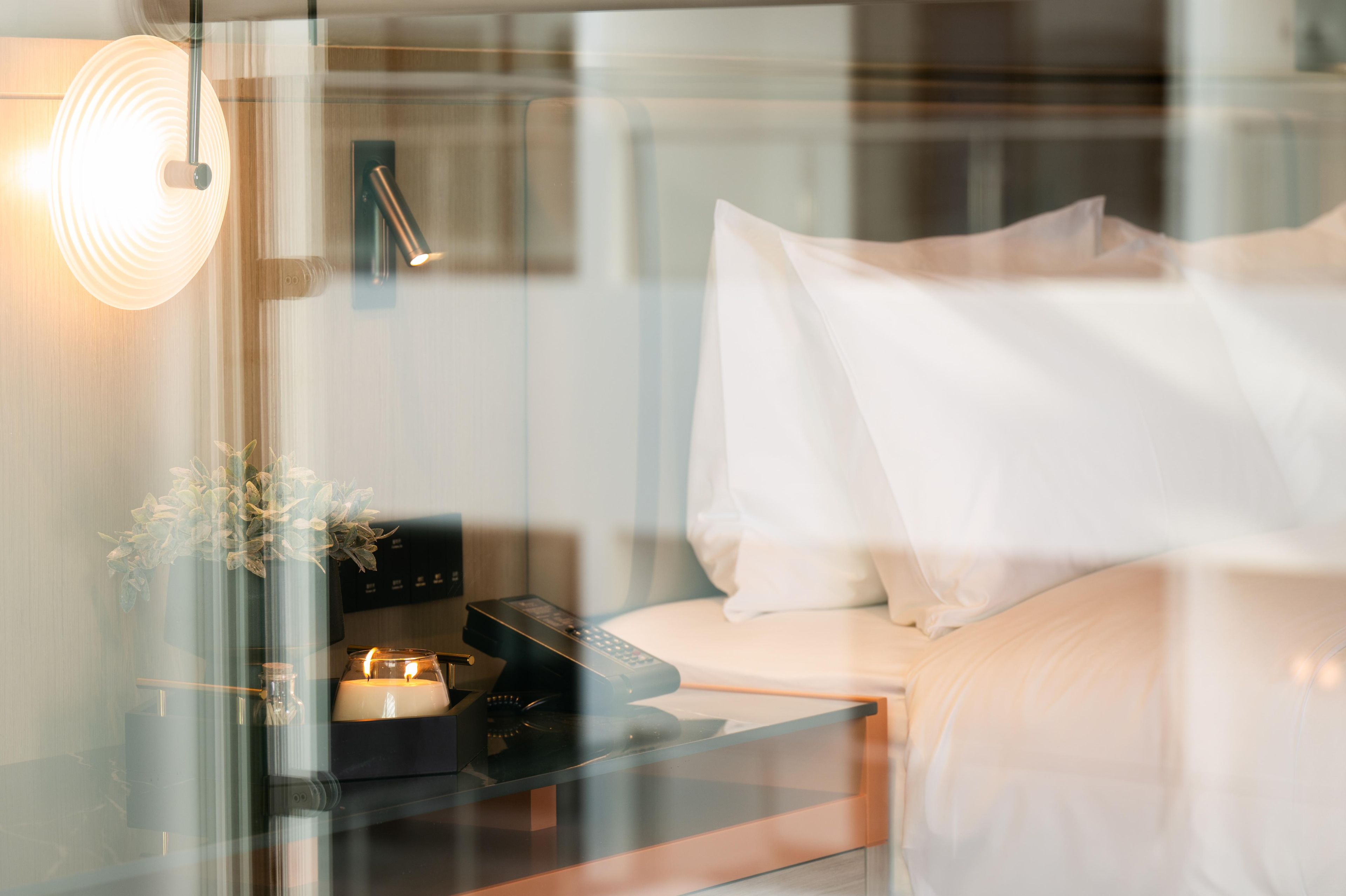 Camera d'albergo moderna con parete divisoria in vetro, comodino con telecomando, candela e vaso con fiori.