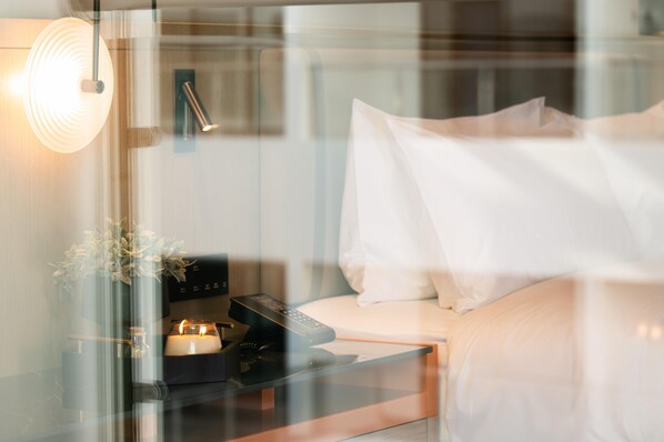 Una habitación de hotel moderna con un separador de vidrio, una mesita de noche con un control remoto, una vela y un florero con flores.