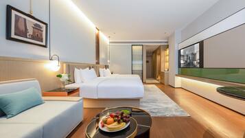 A modern hotel room with a bed, a sofa, a dining table with fruit, and a TV.