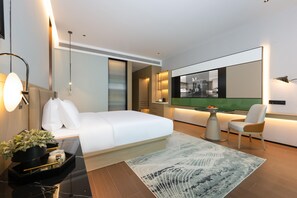 Habitación de hotel moderna con una cama grande, un escritorio con ordenador, una silla, una pequeña mesa con fruta y un espejo grande.