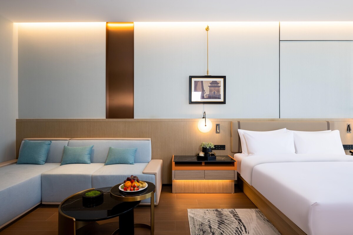 A modern hotel room with a sofa, a bed, a small table with fruit, and a wall-mounted picture.