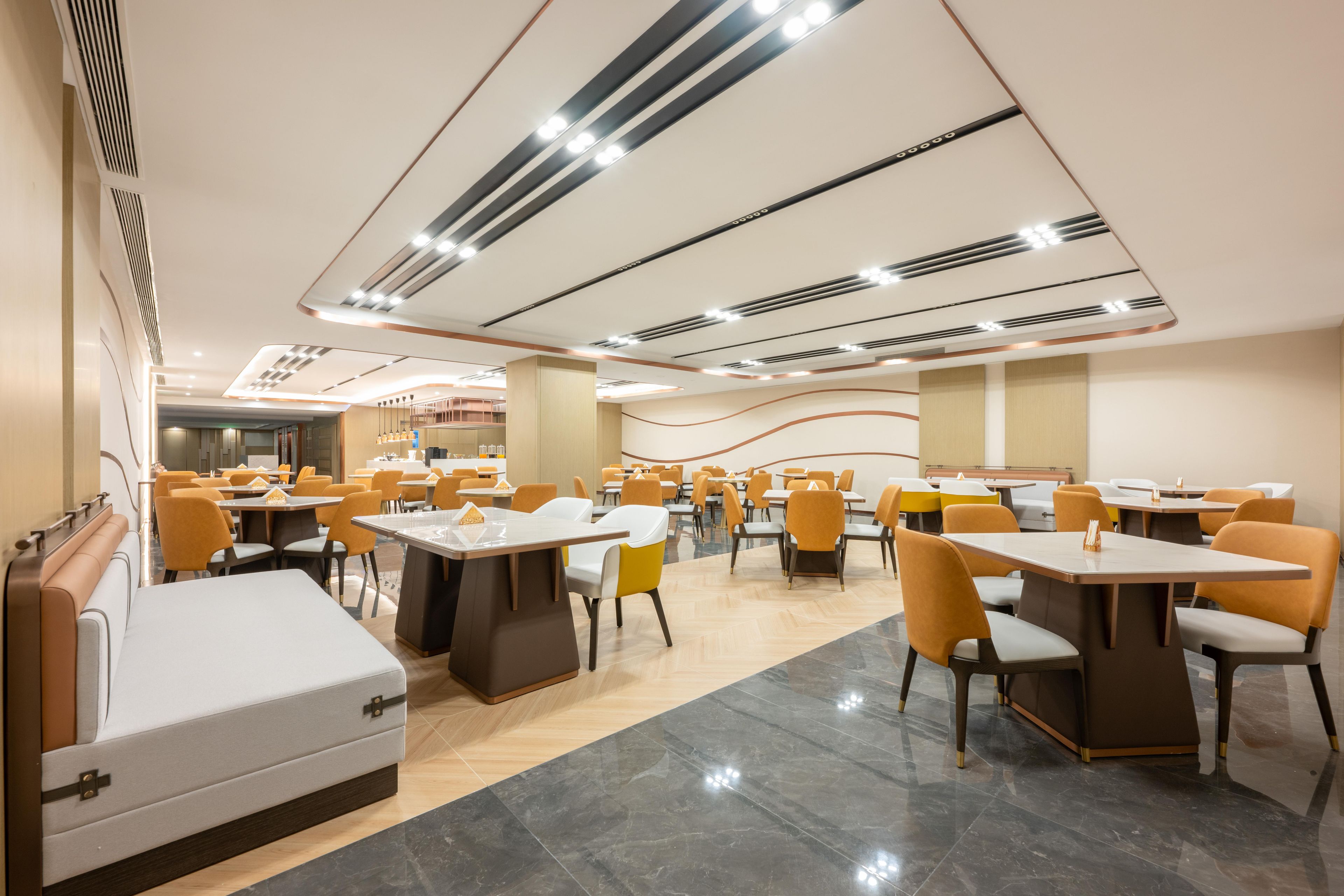 A modern dining area with multiple tables and chairs, a central seating area with cushioned benches, and a ceiling with linear light fixtures.