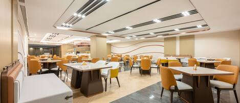 A modern dining area with multiple tables and chairs, a central seating area with cushioned benches, and a ceiling with linear light fixtures.
