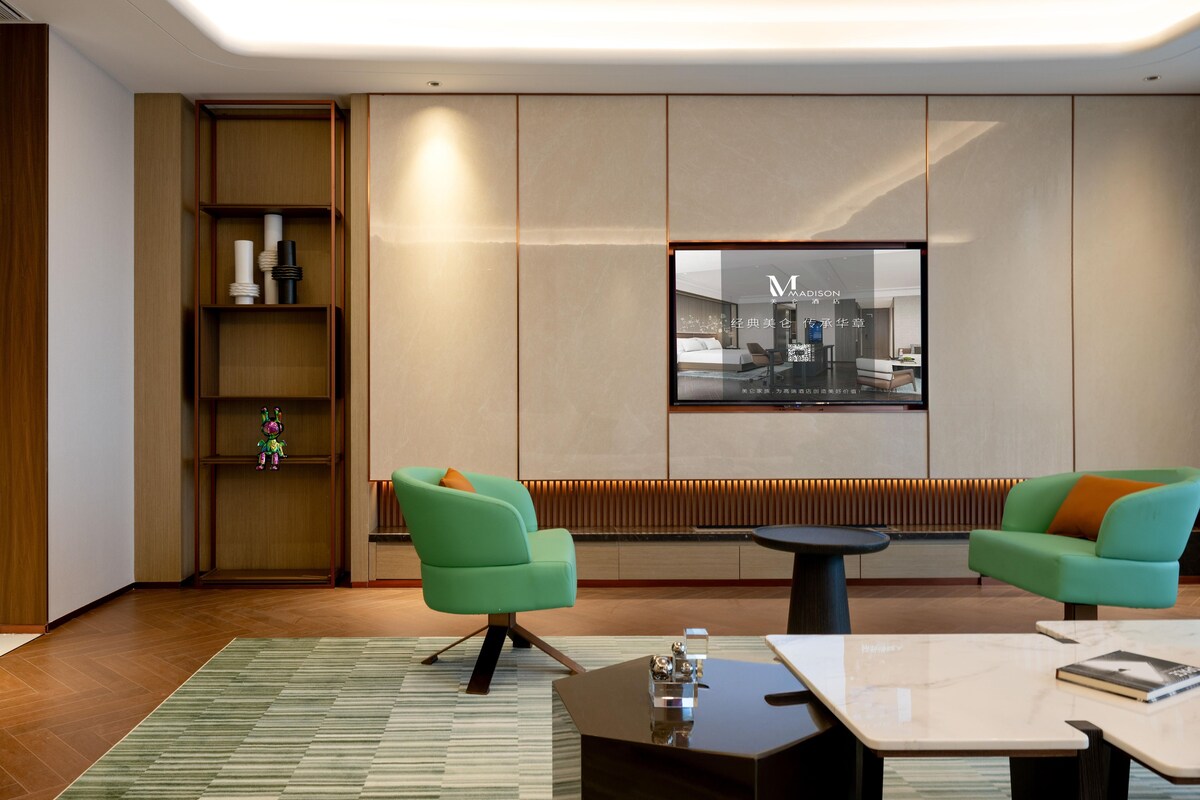 Un moderno vestíbulo de hotel con dos sillones verdes, una pequeña mesa redonda, una estantería y un gran ventanal con vistas a la ciudad.