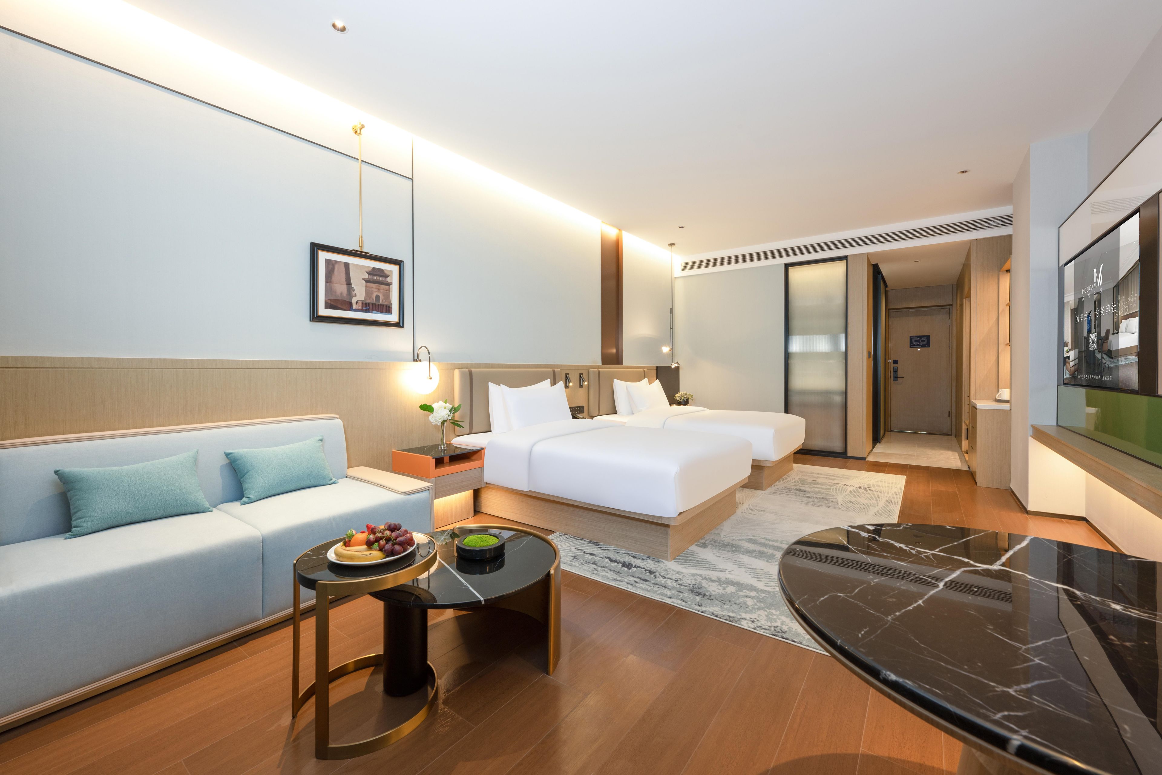 Una habitación de hotel moderna con sofá, cama, mesa de comedor con fruta y un espejo grande.