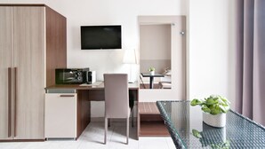 A compact hotel room with a kitchenette, a flat-screen TV, a desk with a chair, a small table with a plant, and a glass partition.