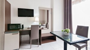 A hotel room with a desk, a flat-screen TV mounted on the wall, a small table with a potted plant, and a window with curtains.
