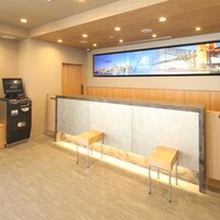 Zona de recepción de un hotel con mostrador, dos bancos de asiento, una pantalla digital y una pantalla en la pared.