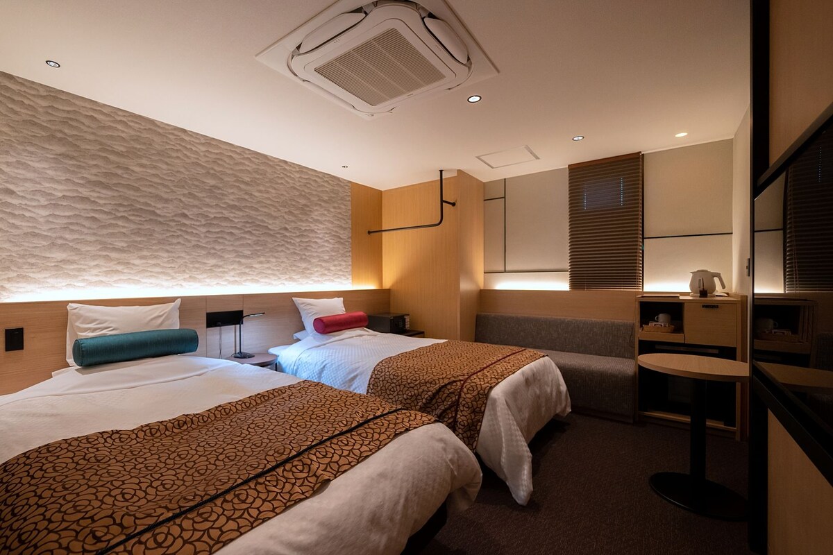 A hotel room with two beds, a small table, a mirror, and a wall-mounted heater.