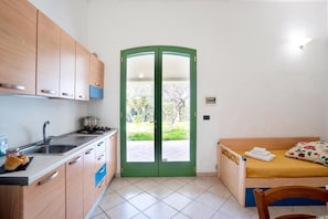 Cocina compacta con pileta, estufa y placares, una cama individual con colchón amarillo, y una puerta verde que da al exterior.