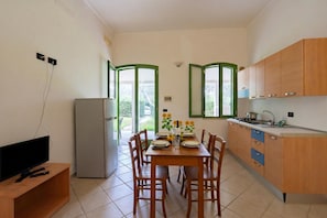A compact kitchen with a dining table, chairs, a refrigerator, and a TV on a wooden stand.