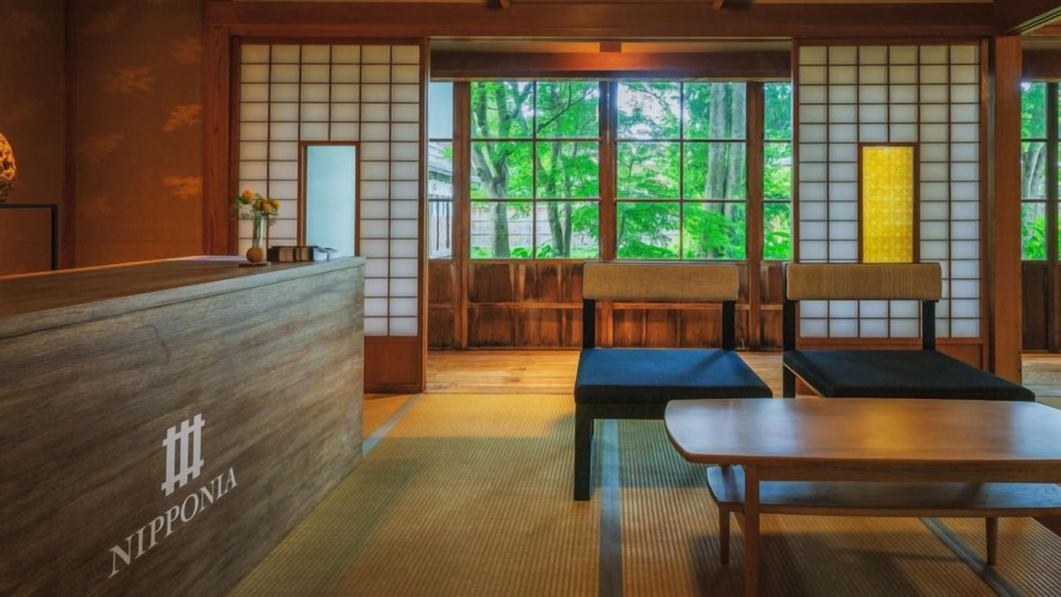 Interior de una posada japonesa tradicional, con suelo de tatami, un mostrador de recepción y vistas a zonas verdes a través de amplios ventanales.