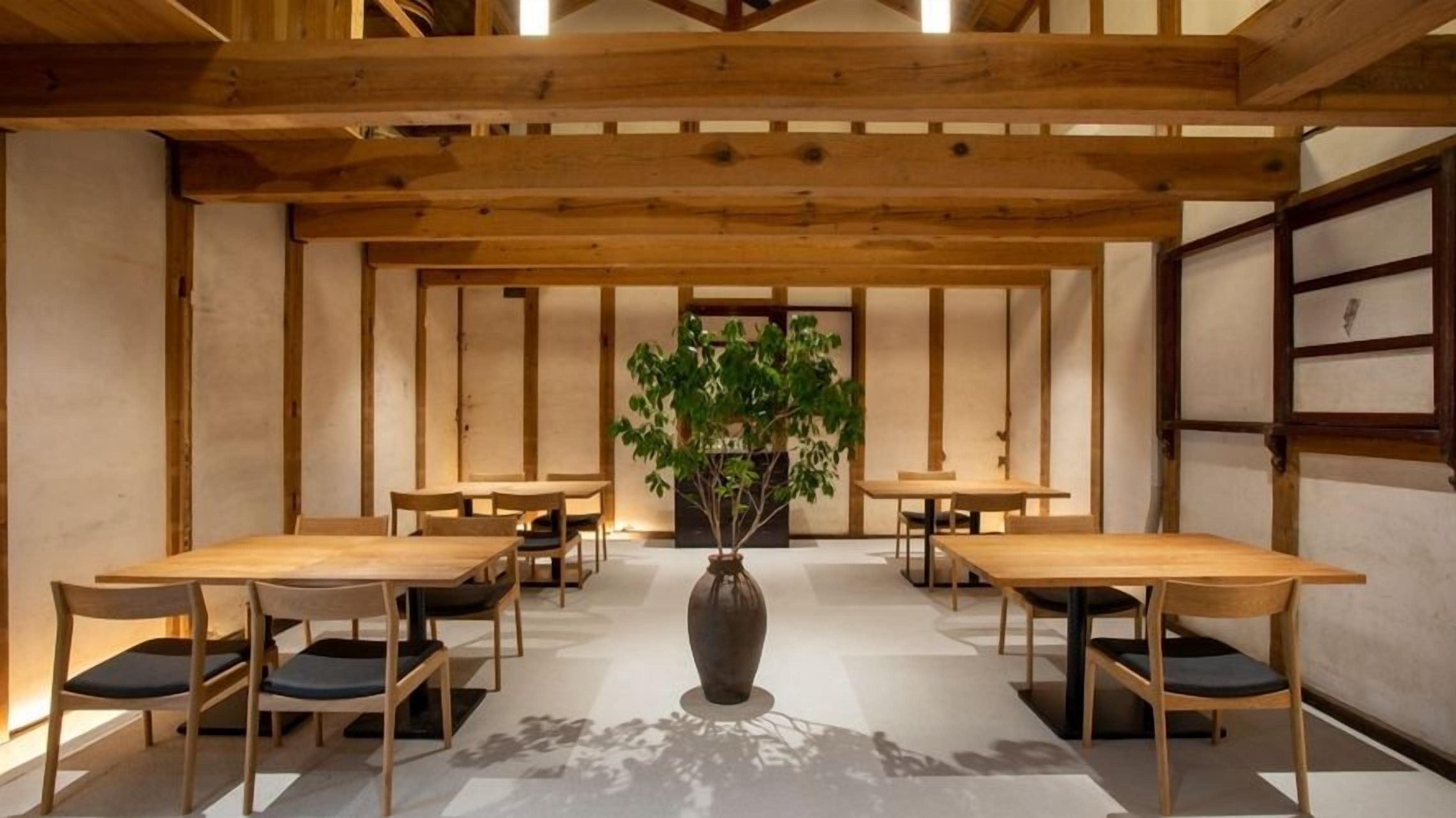 Una habitación con mesas y sillas de madera dispuestas en forma de U, un jarrón grande con una planta en el centro y una decoración de estilo tradicional japonés.
