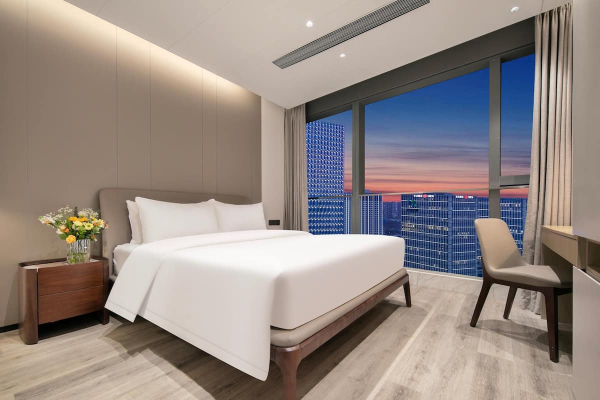 Ein modernes Hotelzimmer mit einem großen Bett, einem Schreibtisch und Blick auf eine Stadtlandschaft bei Sonnenuntergang.
