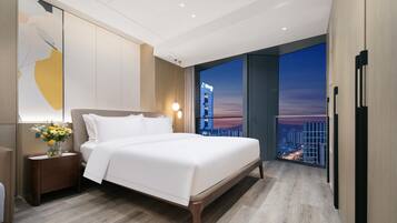 A modern hotel room with a large bed, a nightstand with a vase of flowers, and a city view through large windows.