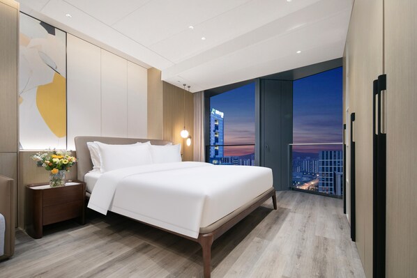 Een moderne hotelkamer met een groot bed, een nachtkastje met een bloevas en een stadsgezicht door grote ramen.