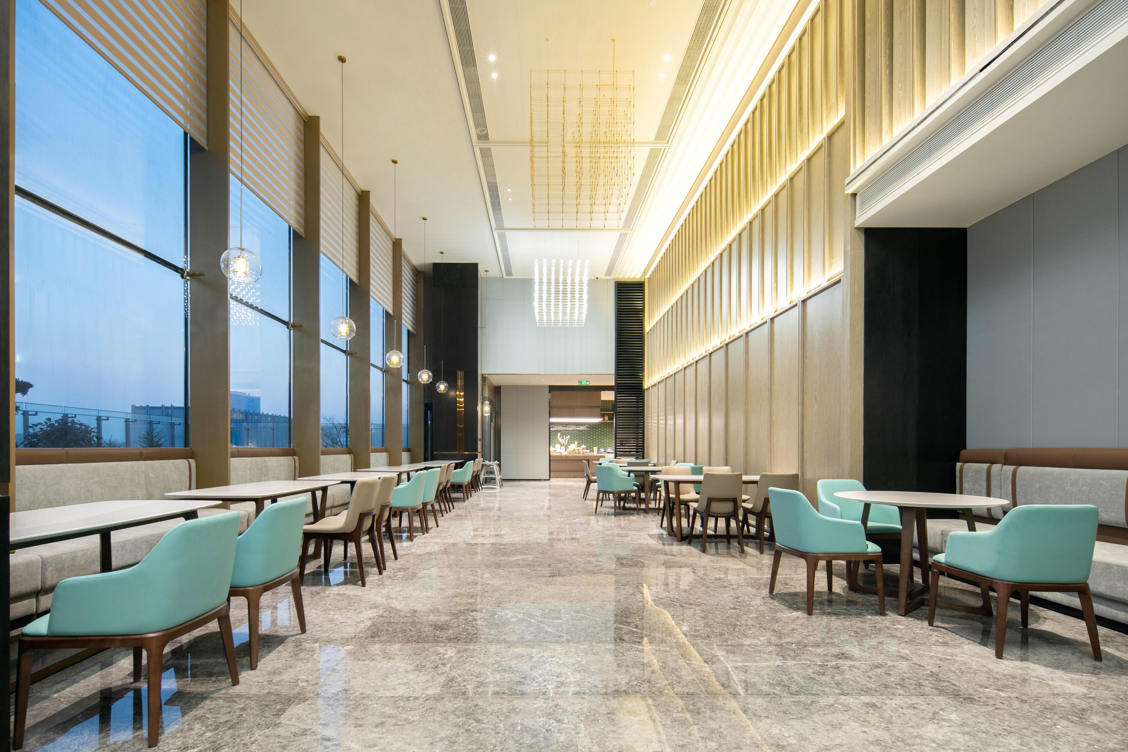 A spacious indoor area with long tables and chairs, large windows, and a modern ceiling design.
