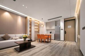A modern living room with a sofa, coffee table, and dining area with chairs. There is a kitchen with a refrigerator and a wooden door.