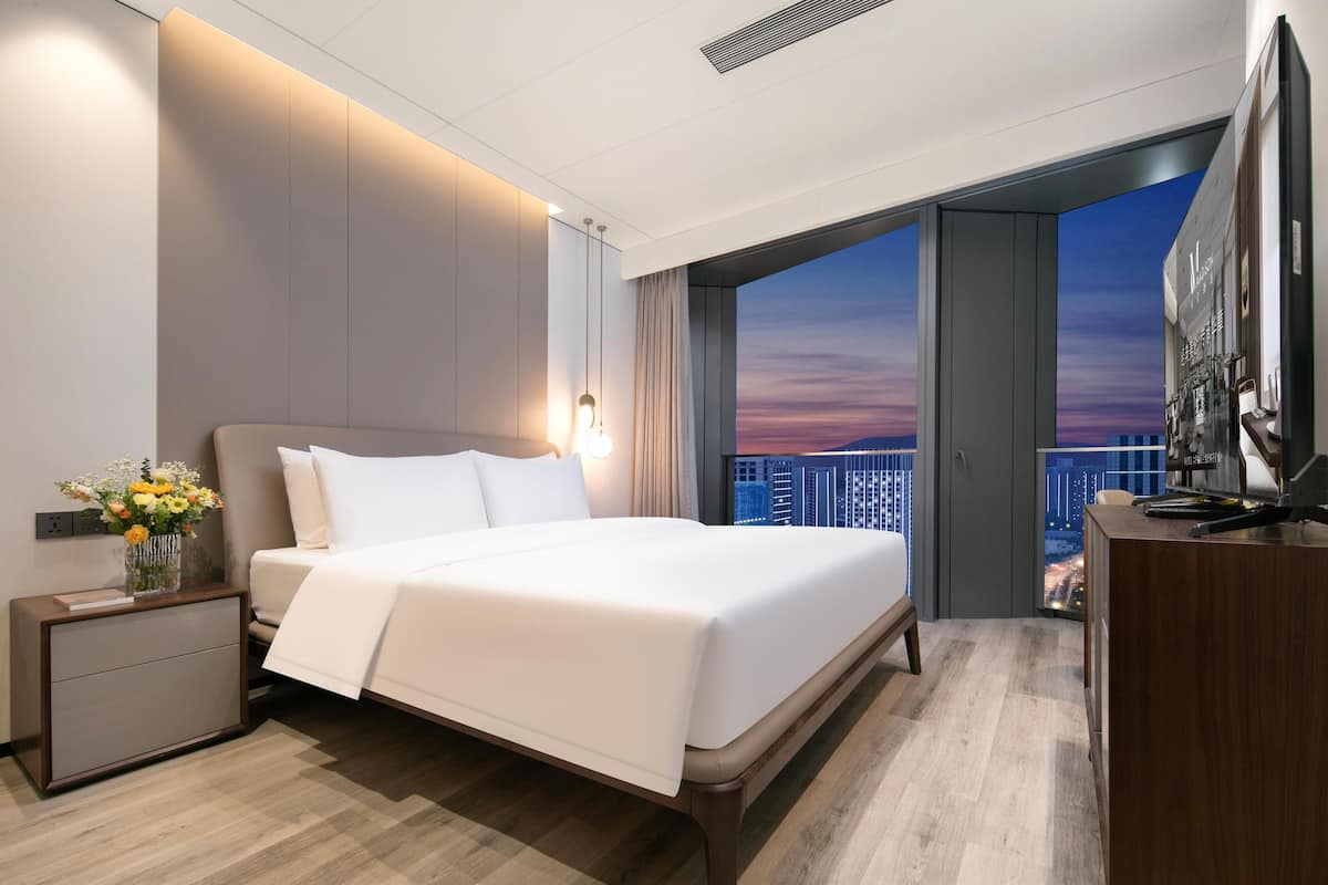 Ein modernes Hotelzimmer mit einem großen Bett, einem Schreibtisch und Blick auf die Stadt durch eine offene Tür.