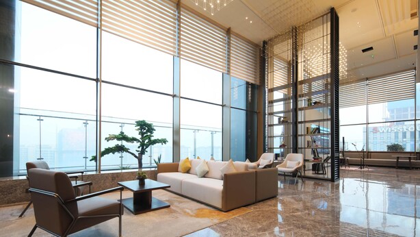 Um lobby amplo com um sofá moderno, poltronas e uma mesa de centro. Grandes janelas proporcionam iluminação natural e vista para a cidade.