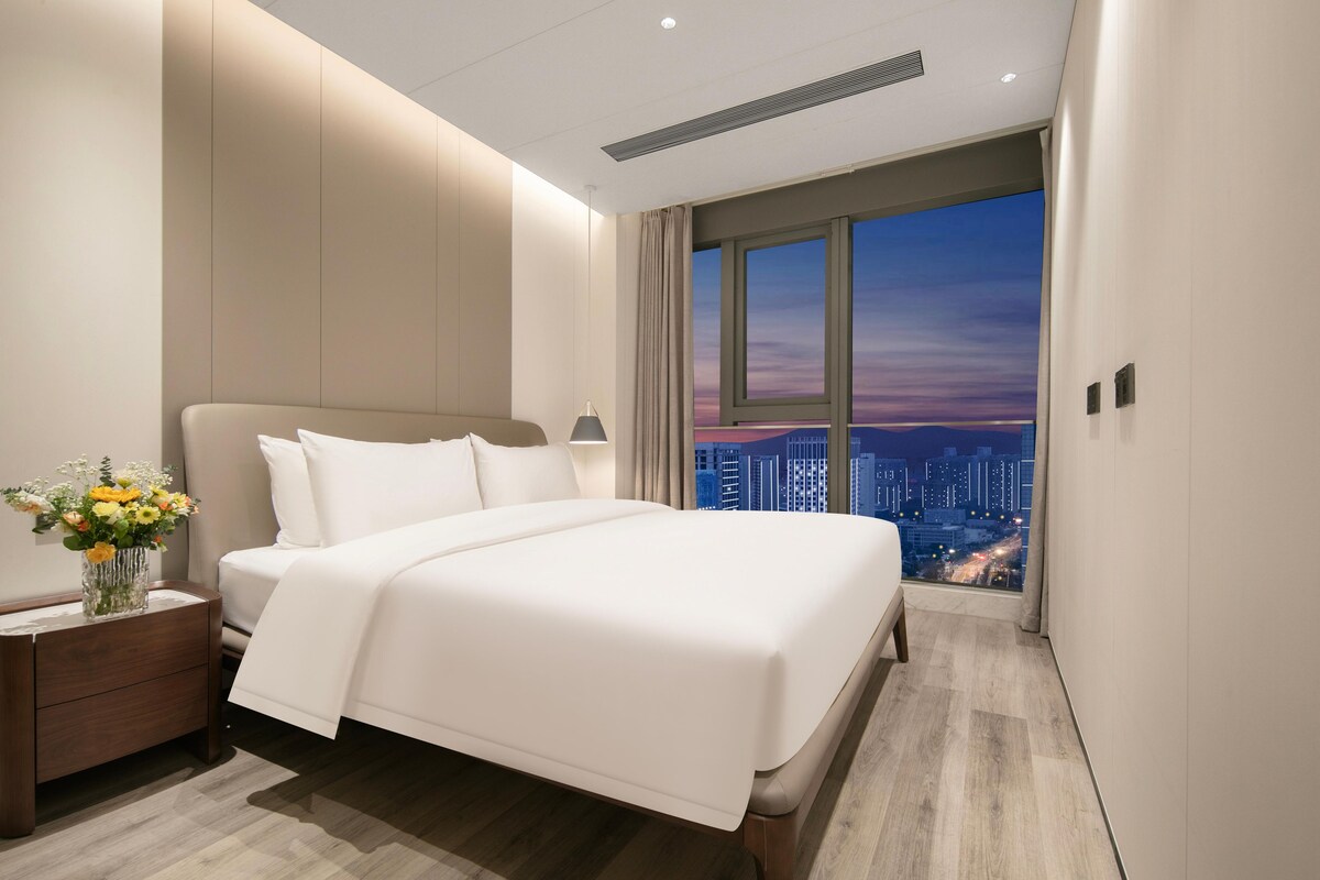 Ein modernes Hotelzimmer mit einem großen Bett, einem Nachttisch mit Blumen und Blick auf die Stadt durch das Fenster.