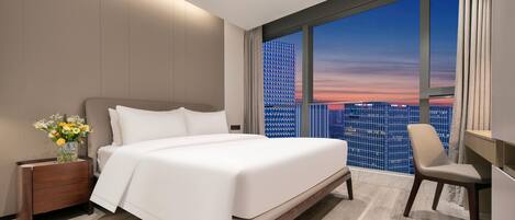 A modern hotel room with a large bed, a desk, and a view of a cityscape at sunset.