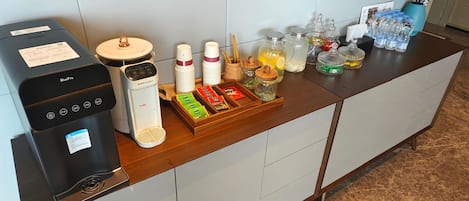 Zona de recepción de hotel con una máquina de café, vasos desechables, una bandeja con bolsitas de té, sobres de azúcar y un jarrón con flores.
