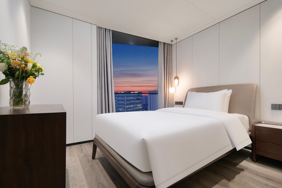 Een moderne hotelkamer met een groot bed, een nachtkastje, een bloemenvaas en uitzicht op de skyline van de stad door een groot raam.