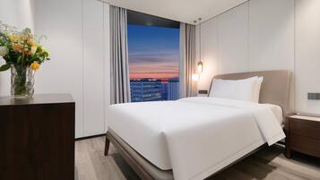 A modern hotel room with a large bed, a nightstand, a vase of flowers, and a view of the city skyline through a large window.