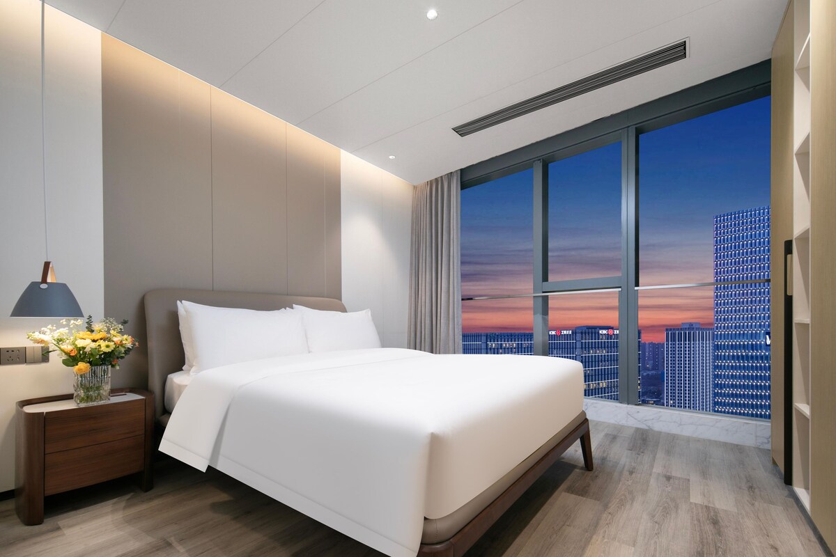 Une chambre d’hôtel moderne dotée d’un grand lit, d’une table de chevet avec une lampe et offrant une vue sur le paysage urbain à travers de grandes fenêtres.