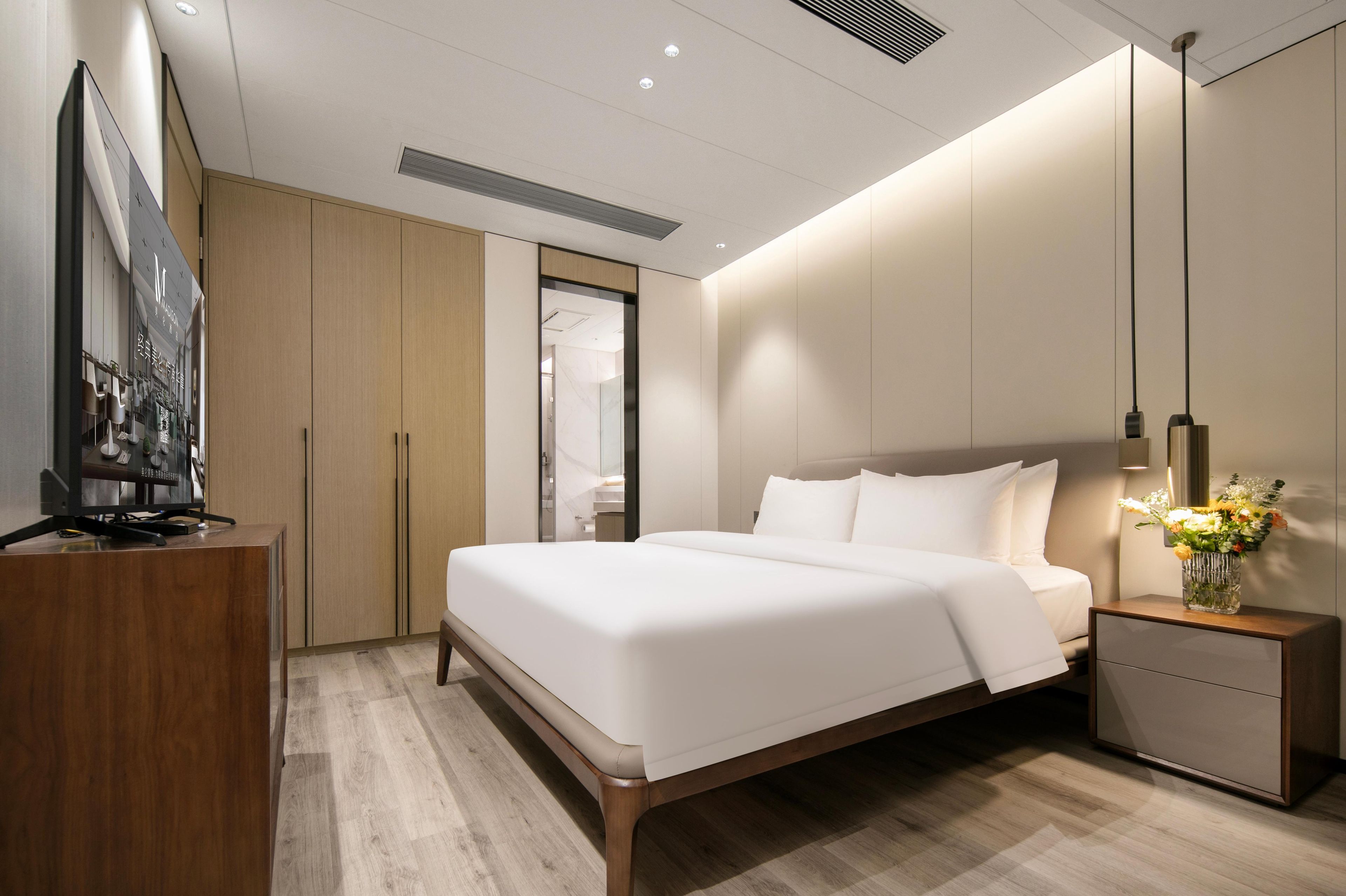 A modern hotel room with a large bed, a wooden desk, a TV, and a nightstand with a vase of flowers.