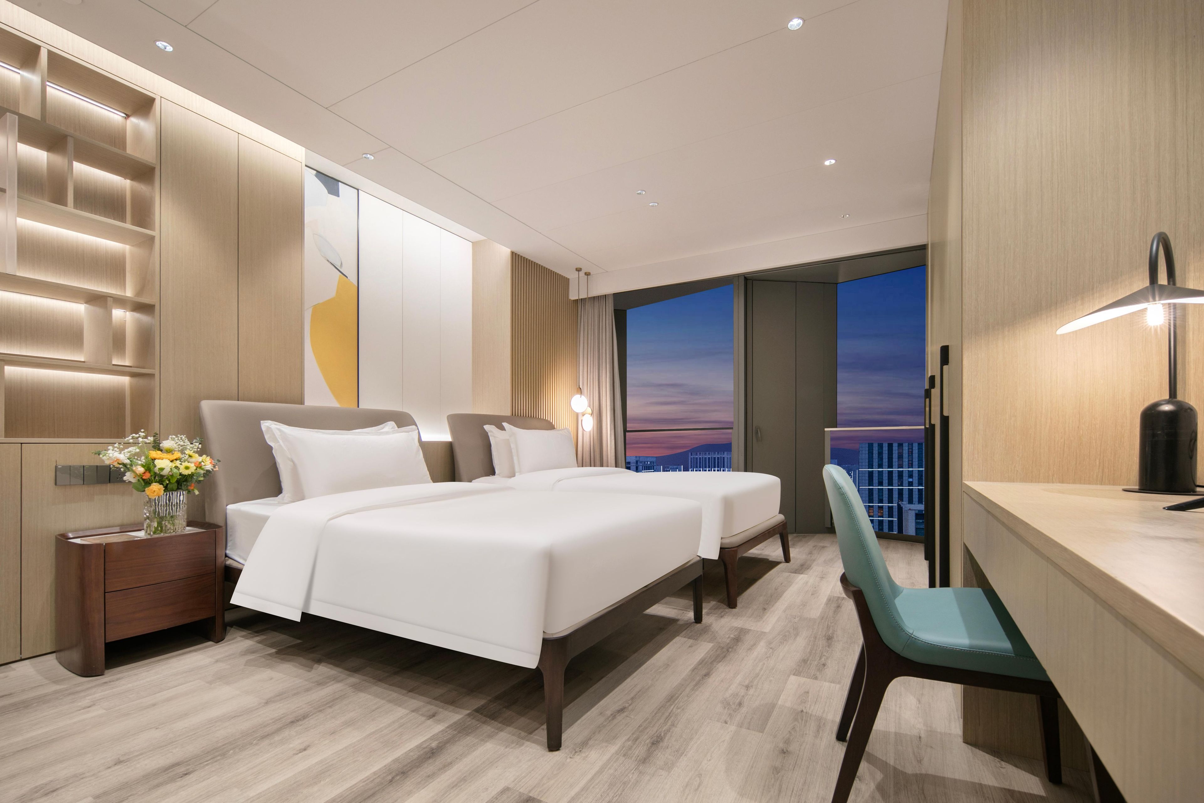 Ein modernes Hotelzimmer mit einem großen Bett, einem Schreibtisch und Blick auf die Stadt bei Sonnenuntergang.