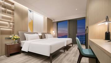 A modern hotel room with a large bed, a desk, and a view of the city at sunset.