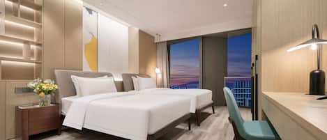 A modern hotel room with a large bed, a desk, and a view of the city at sunset.
