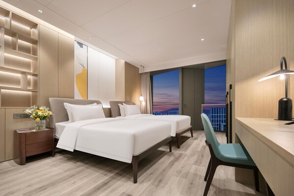 A modern hotel room with a large bed, a desk, and a view of the city at sunset.