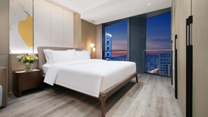 Quarto de hotel moderno com uma cama grande, uma mesa de cabeceira com um vaso de flores e vista para a cidade através de amplas janelas.