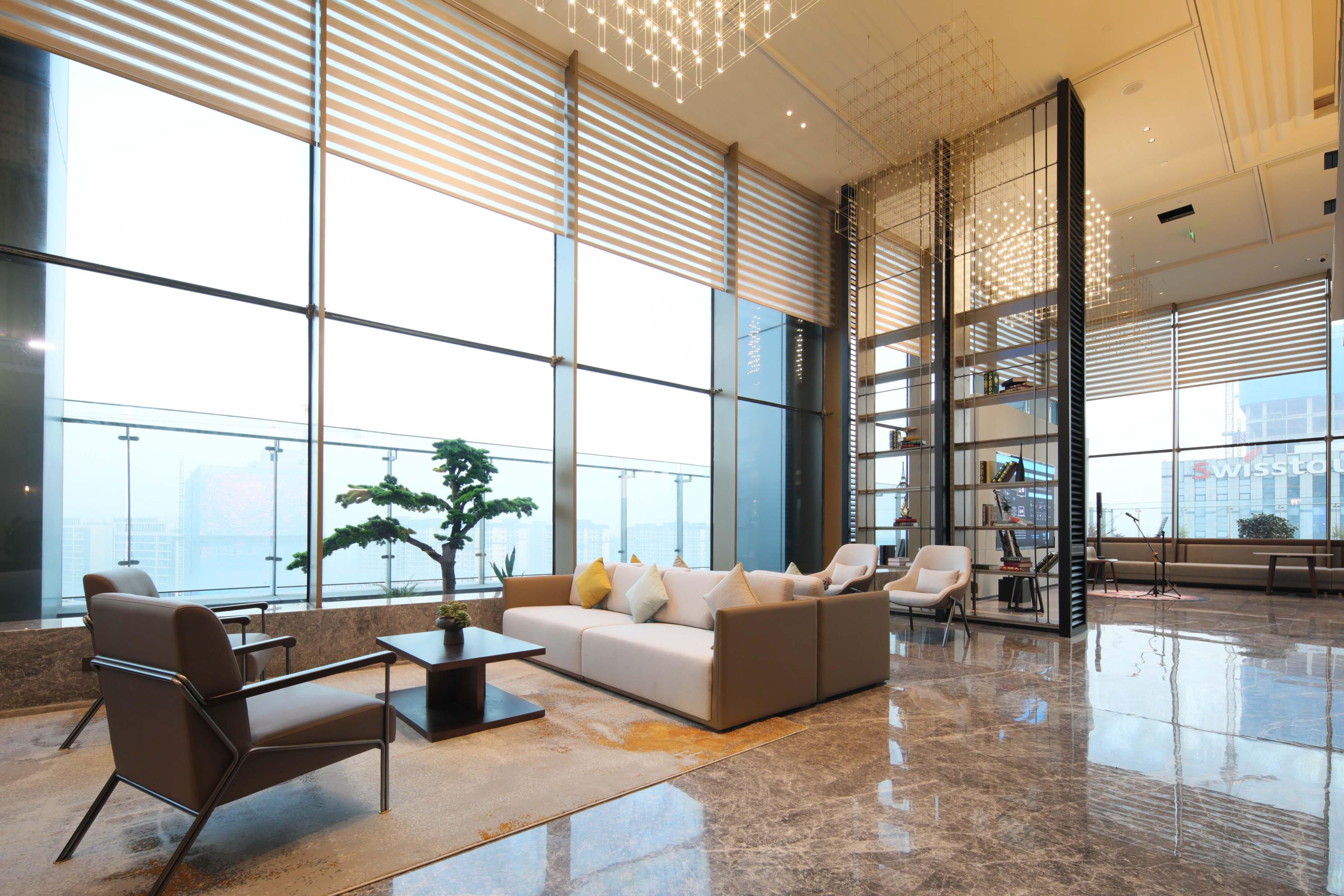 A spacious lobby with a modern sofa, armchairs, and a coffee table. Large windows provide natural light and a view of the city.