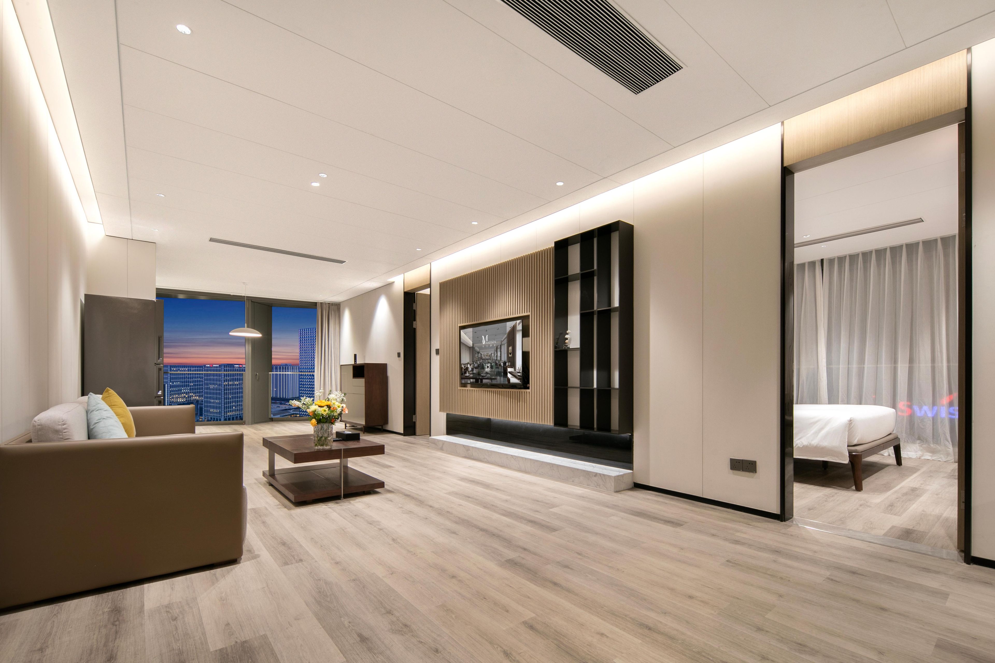 Habitación de hotel moderna con una cama grande, una zona de estar con sofá y mesa de centro, un televisor montado en la pared y vistas a la ciudad a través de la ventana.